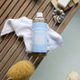 A bottle of Dr. Bronners Pure-Castile Liquid Soap rests on a white towel on wooden slats, with a yellow sponge, natural soaps, and a baby teething toy nearby.
