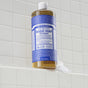 A bottle of Dr. Bronner’s Pure-Castile Liquid Soap rests on a white tiled ledge, suds spilling over. This versatile vegan soap features a blue label with white text and is renowned for its many castile soap uses.