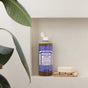 A bottle of Dr. Bronners Pure-Castile Liquid Soap stands on a white recessed shelf beside a bar of vegan soap, a scrub brush, bath sponge, and a leafy green plant partially visible on the left.