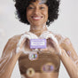 A smiling woman with curly hair, covered in bubbles, holds Dr. Bronner’s Pure-Castile Bar Soap in front of her chest. More soap bubbles float around her against a light background.