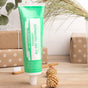 A green tube of Dr. Bronner’s All-One Toothpaste stands upright on a wooden surface beside a toothbrush, wrapped gifts, and a small straw ornament, with grasses and white tiles in the background.