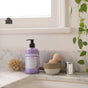 A bottle of Dr. Bronners Organic Sugar Soap rests on a marble counter beside a wooden brush, a bowl with a wooden item, a soap dispenser, and a hanging vine—an inviting scene featuring sustainable packaging.