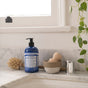 A blue bottle of Dr. Bronners Organic Sugar Soap rests on a marble countertop near a dish brush, bowl with wooden scoop, small metal cup, and leafy plant by the window; this soap is vegan and cruelty-free.