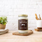 A jar of Dr. Bronners Whole Kernel Virgin Coconut Oil sits on a wooden board in a bright kitchen with a white brick wall, surrounded by a potted plant, towel, and utensils—a taste of Sri Lanka’s best.