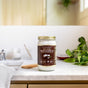 A jar of Dr. Bronners Whole Kernel Virgin Coconut Oil sits on a marble bathroom counter beside a wooden comb, with green plants and a blurred sink in the background.