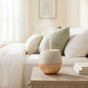 The Drift Linen Diffuser by Plant Therapy releases a fine mist and soothing ambient light while sitting on a wooden nightstand next to stacked books, with a cozy bed filled with soft pillows in the background.