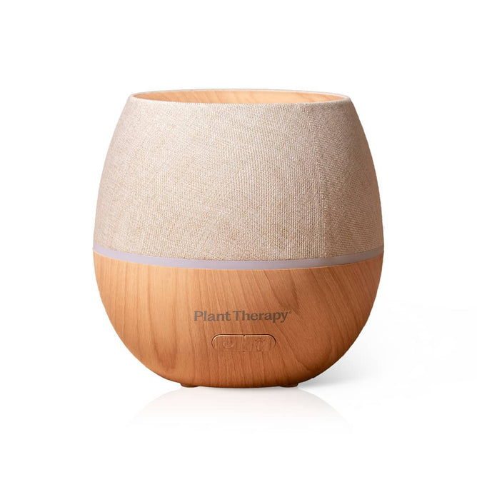 The Drift Linen Diffuser by Plant Therapy features a round wood-grain design with a beige fabric top, an ambient light feature, and the Plant Therapy logo on the front.