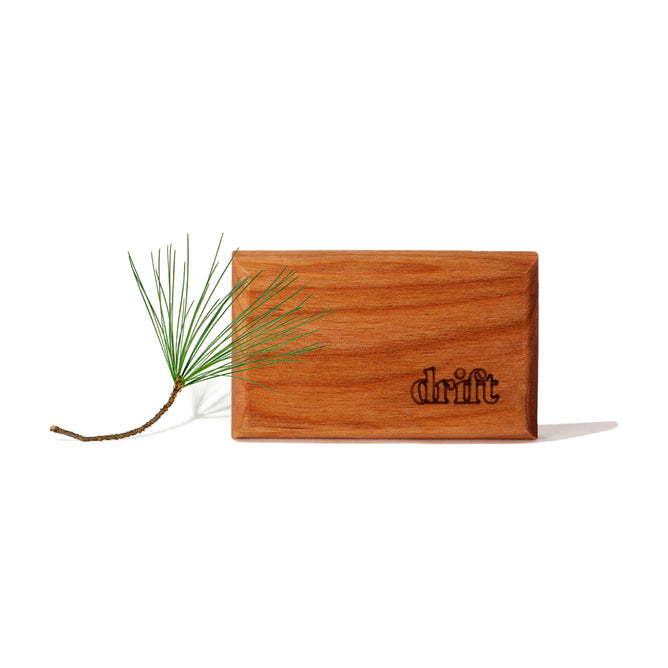 A small pine twig lies beside a rectangular wooden block engraved with drift, evoking the Drift Wood Car Air Freshener Refill by Drift, which delivers premium scents for your space, all set against a white background.