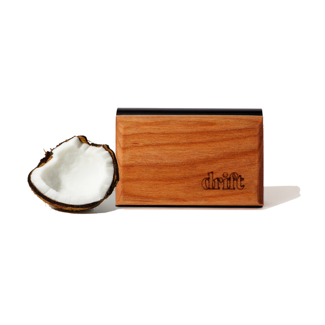 The Drift Single Wood Car Air Freshener Starter Kit, crafted from sustainably sourced wood and engraved with Drift, is shown beside a halved coconut on a plain white background, highlighting its natural inspiration.