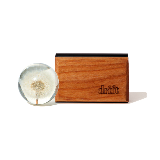 A clear resin sphere with a dandelion puff rests beside the Drift Single Wood Car Air Freshener Starter Kit, which features sustainably sourced wood and an engraved Drift logo, displayed on a white background.