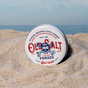 A round tin of Old Salt Pomade, featuring a skull and anchor on the lid, rests on sandy beach with ocean and sky in the background.