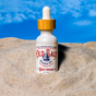 A bottle of Old Salt Beard Oil (Sea Kelp) with a wooden dropper cap stands upright in sand under a bright blue sky. The label features nautical-themed graphics and reads Sea Kelp.