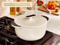The Caraway Cast Iron Dutch Oven with Flat Lid, 6.5 qt, sits on a stove in a kitchen with fresh tomatoes, olive oil, and a cutting board nearby. This non-toxic cookware offers easy cooking and cleaning plus superior heat retention with durable enamel.