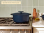 A Caraway Cast Iron Dutch Oven with Flat Lid, 6.5 qt, in navy blue rests on a gas stove by a white countertop holding a terra-cotta bottle and oil dispenser. A cleaning tip box is displayed on the tiled wall, highlighting its heat retention.