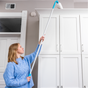 A woman in a blue sweater uses the E-Cloth Easy-Click Home Cleaning Set with reusable pads to clean the top of a tall white cabinet in a bright room.