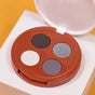 The Coffee Oil Infused Pigmented Eyeshadow Palette by Everyday For Future features black, silver, light beige, and dark gray shades in a circular brown compact, photographed on a white and yellow background.