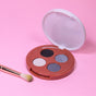 The Everyday For Future Coffee Oil Infused Pigmented Eyeshadow Palette, with black, silver, beige, and blue shades and a white lid, sits open on a pink surface next to a makeup brush.