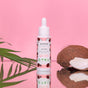 A dropper bottle of Everyday For Future’s Moisturizing Face Serum with Hyaluronic Acid stands upright on a pink background, flanked by a green palm leaf and halved coconut, highlighting its hydrating benefits and tropical-inspired design.