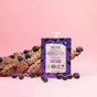 A pouch of Firming Blueberry Face Scrub by Everyday For Future sits amidst scattered blueberries and tree bark on a pink backdrop. The package emphasizes its antioxidant and vegan features, appealing to eco-conscious skincare fans seeking a burst of berry goodness.