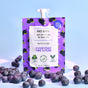 A sachet of Firming Blueberry Face Scrub labeled Berrylicious, with scattered blueberries, highlights its vegan formula and antioxidant properties. The purple packaging features black blueberry illustrations and emphasizes Everyday For Future and Cruelty-Free against a soft blue background.