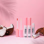 Three pink tubes of Everyday For Future Moisturizing & Non-Sticky Coconut Lip Oil, one opened to show the applicator, are arranged on a pink background with coconut halves, emphasizing the nourishing coconut oil infusion and soft shadows.