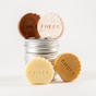 The Complete Minibar Set by Ember Wellness displays four EMBER-embossed round moisturizing bars in brown, pale pink, yellow, and gold, arranged around two stacked metal tins on a clean white background.