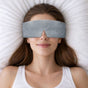 A woman with long brown hair relaxes on a white pillow, smiling gently while wearing the Elseware Unplug Plush Eye Mask and a white tank top.