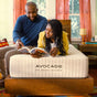 A man and woman read a book together while lying on an Eco Organic Mattress by Avocado Green Mattress in a cozy, sunlit bedroom.