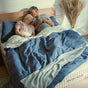 An older couple relaxes in bed together, the woman resting her head on the man’s shoulder while he holds a mug. The bedding is blue and white, and Avocado Green Mattress is visible on the mattress—an Eco Organic Mattress that is GOTS-certified to ensure their bedroom is both comfortable and nontoxic.