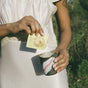 A person in a light pink dress pours a Lemon Ginger Immune Toddy - Sachets by Wooden Spoon Herbs into a striped ceramic cup, with greenery and sunlight brightening the background.