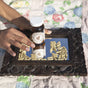 A hand arranges a bottle labeled “Wooden Spoon Herbs Oregano Daily Wellness Capsules” and green capsules—herbal immune support—on a carved wooden tray with a mirror, set on a floral quilt. Both the bottle and capsules are reflected in the mirror.