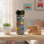 An 18oz Elemental Stainless Steel Kids Water Bottle with a pop fidget handle and yellow construction vehicles sits on a light wood table beside a potted succulent and toy truck. A bed and framed car pictures are blurred in the background.