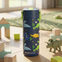 An Elemental Stainless Steel Kids Water Bottle, 18oz with a blue pop fidget handle and colorful dinosaur design sits among toy blocks and a triceratops on a wooden floor, sunlight shining through the window behind.