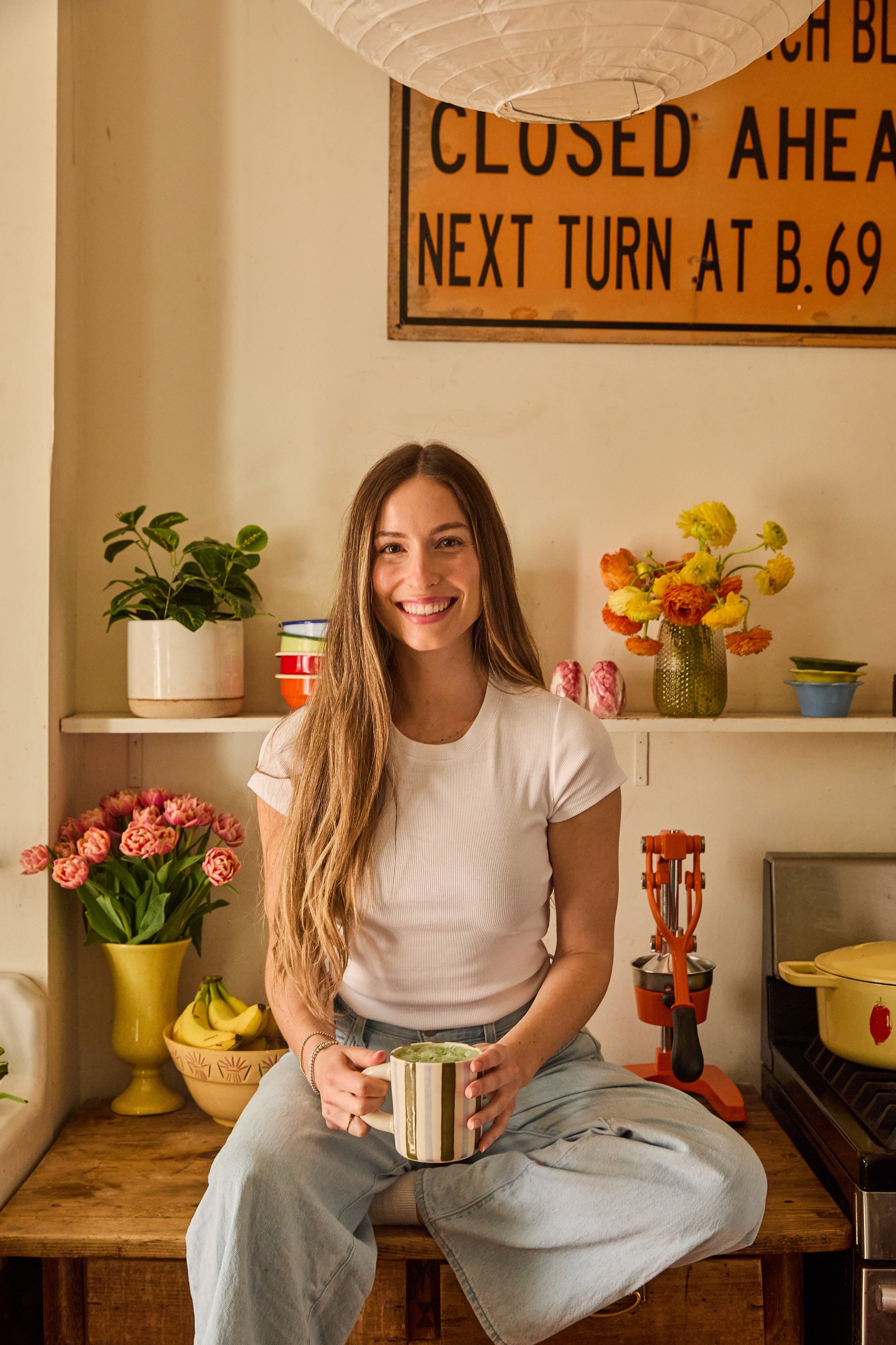 A woman with long brown hair, wearing a white t-shirt and light blue jeans, sits smiling on a kitchen counter holding a mug. She is surrounded by colorful flowers and potted plants, with a vintage sign on the wall behind her.