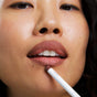 A close-up of a person with a nose ring, natural textured skin, and slightly parted lips holding Ere Perez’s Acai Lip Pencil, a nude shade, to their lips.