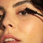 A close-up shows someone applying Ere Perez Mini Avocado Mascara to their upper lashes, highlighting a natural makeup look. Their smooth, dewy skin and freckles on the cheek and nose enhance the eye-focused shot.