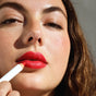 A close-up shows a woman with fair skin and brown hair applying Ere Perez Coco Crayon Lipstick to her lips, holding it in her right hand. Her dewy skin, natural makeup, and defined brows complete an effortlessly fresh look.