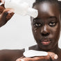 A woman with dark skin and natural makeup pours Ere Perez Ginkgo Micellar Cleanser from a white bottle onto a cotton pad, wearing a white tank top against a light background.