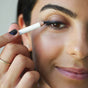 A close-up of a woman applying Ere Perez Jojoba Eye Pencil to her upper eyelid. Her well-groomed brows and natural makeup highlight the gentle formula, ideal for sensitive eyes. She smiles slightly and wears a thin gold ring.