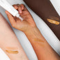 Three forearms with varying skin tones display swatches of Ere Perez Peach Gel Bronze Drops, a golden shimmering liquid bronzer. A hand holds the white tube, dispensing the sensitive-safe formula onto the middle arm against a white background.