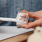 A hand with metallic silver nails holds a white coco crayon makeup pencil inside the Ere Perez Perfect Catch Duo Sharpener, ensuring precise sharpening, with a blurred background.