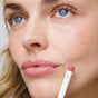 A close-up of a woman’s face with natural makeup, highlighting her blue eyes and pink lips. She holds the Ere Perez Acai Lip Pencil near her lips. Her complexion looks smooth and clear.