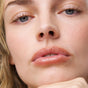 A close-up of a woman highlights her natural skin, blue eyes, and calm expression as her hand rests under her chin, showcasing the smooth finish and lasting color of Ere Perez Acai Lip Pencil on her lips.