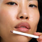 A close-up of a person with smooth skin and a gold nose ring holding the Ere Perez Acai Lip Pencil—a long-lasting, plastic-free white lip liner labeled "pout" and made in Italy—near their lips.