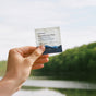 A hand holds a pack of Ursa Major Essential Face Wipes, made with clean ingredients, outdoors with trees and a lake visible in the bright background.