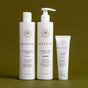 Three white bottles of Innersense Organic Beauty's Essential Hydration Ceremony (shampoo, conditioner, and hair mask) stand upright on an olive green background. The labels display minimalist text and a simple brown floral logo.