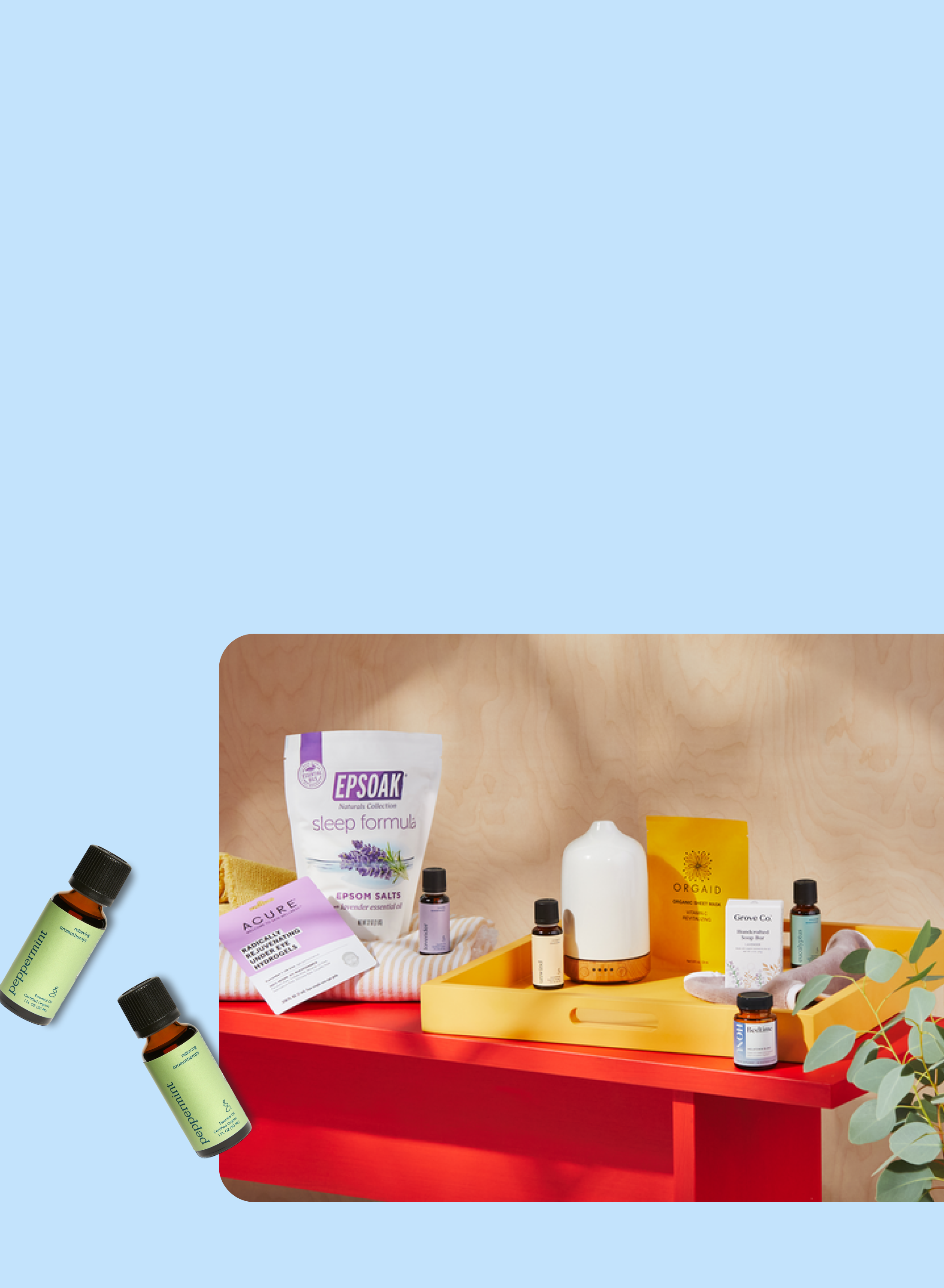A variety of wellness products, including essential oils, bath salts, a white diffuser, a yellow tray, a gold face mask, and a jade roller, are arranged on a red and yellow surface against a light blue and wood background.
