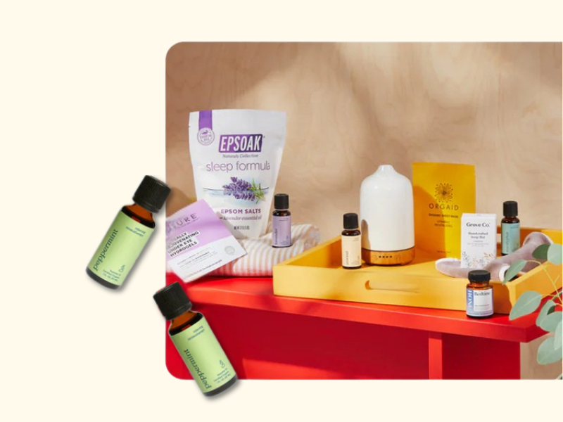 A red table displays essential oils, a white diffuser, Epsom salts, a yellow tray, a striped towel, and wellness products. Two small bottles labeled Peppermint are placed to the left of the table on a cream background.