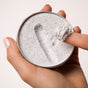 A hand holds an open travel mini tin of Grove Collaborative's The Exfoliating Body Wash Concentrate, revealing the white, speckled cream inside as a finger scoops some out against a plain, light background.