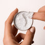 A hand holds an open Grove Collaborative The Exfoliating Body Wash Concentrate Tin, revealing white cream with black exfoliating specks as a finger scoops some out; both manicured nails are in neutral shades against a plain, light background.